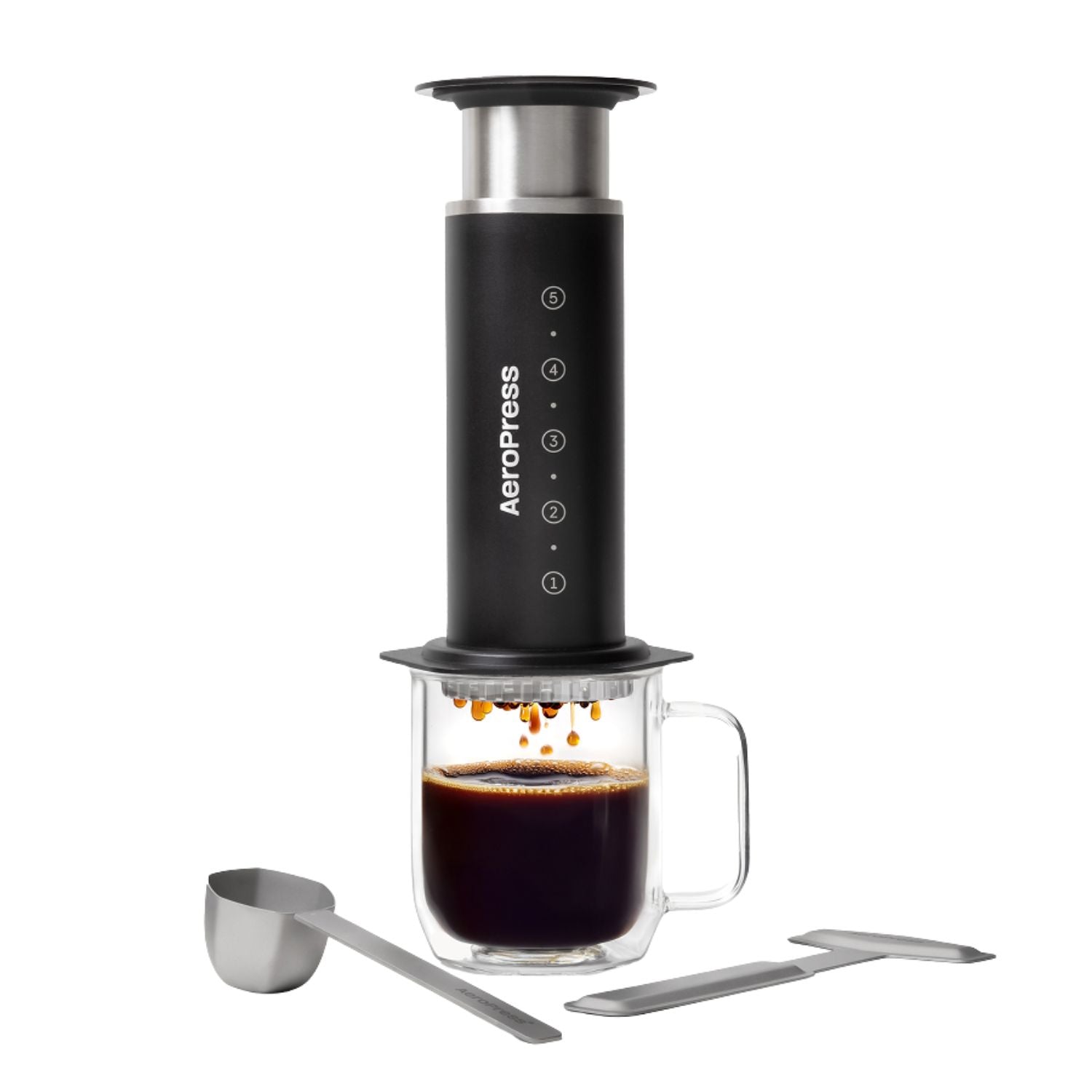 An AeroPress coffee maker with a black body, silver metal top and accessories, shown with a clear glass mug filled with espresso and two metal tools on a white background.